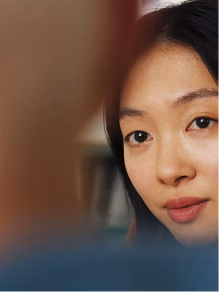 A young woman with dark hair and a thoughtful expression gazes directly at the camera against a blurred background.